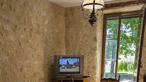 A dining area with a stone wall, a wooden table, chairs, a television on a stand, and a window with a view of greenery. - In a mansion of the nineteenth century, cottage for 5 people (forcalquier)