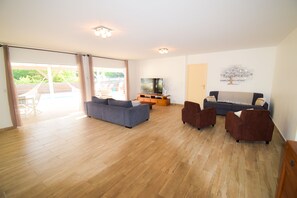 A spacious living room with a large sofa, two armchairs, and a television on a wooden stand. There is a sliding glass door leading to an outdoor area with a table and chairs. - Villa MORENE, near the beach 50m (Sainte-Anne)