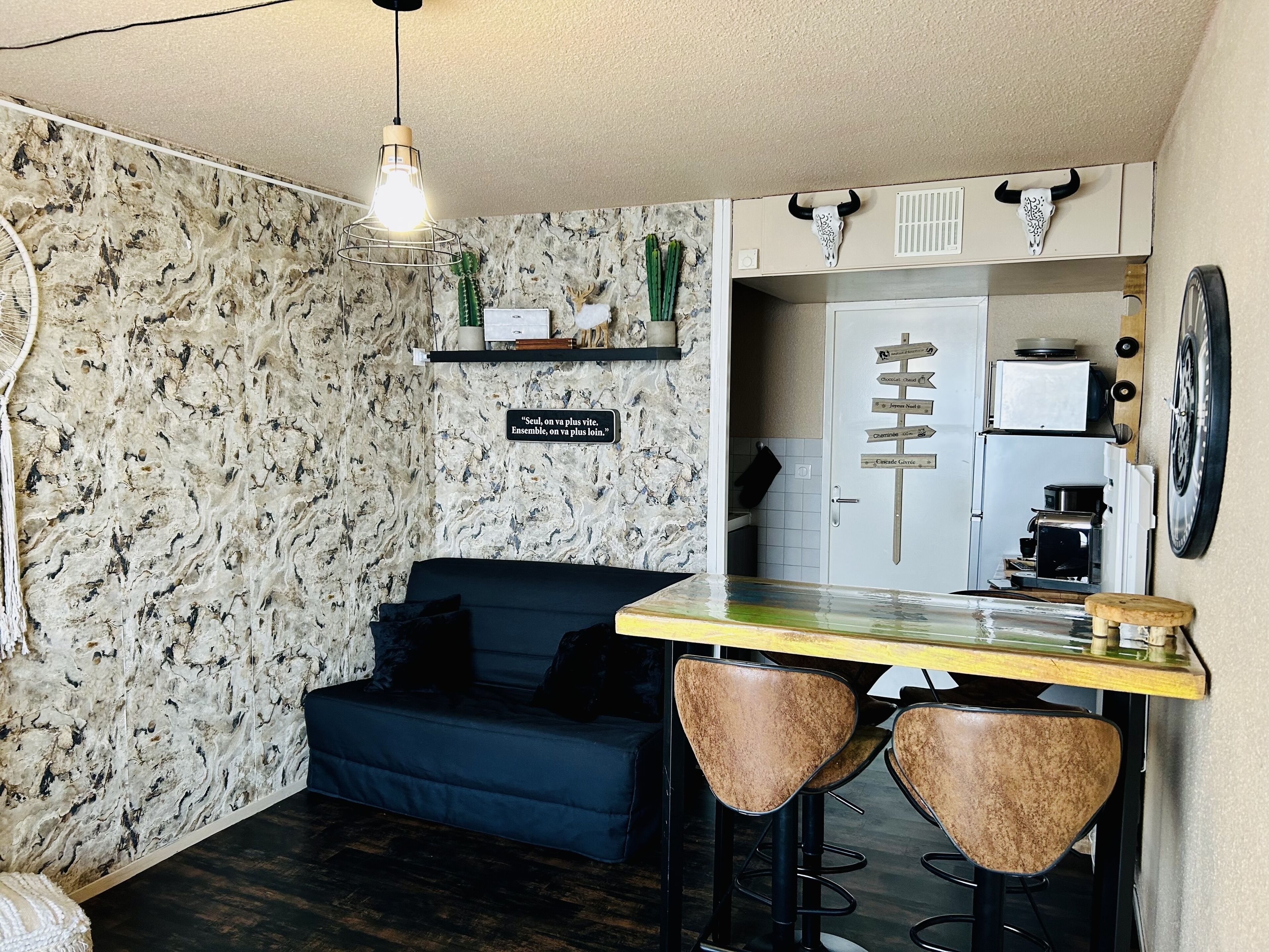 Ein kompakter Wohnbereich mit einer Couch, einer Bar mit Hockern, einem an der Wand befestigten Schild und einer Küchenzeile mit Geräten.