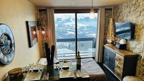 A dining area with a table set for two, a clock, a television, and a view of snow-covered mountains. - Apartment 100% ski in ski out (Les sybelles) 4 people (Le Corbier)