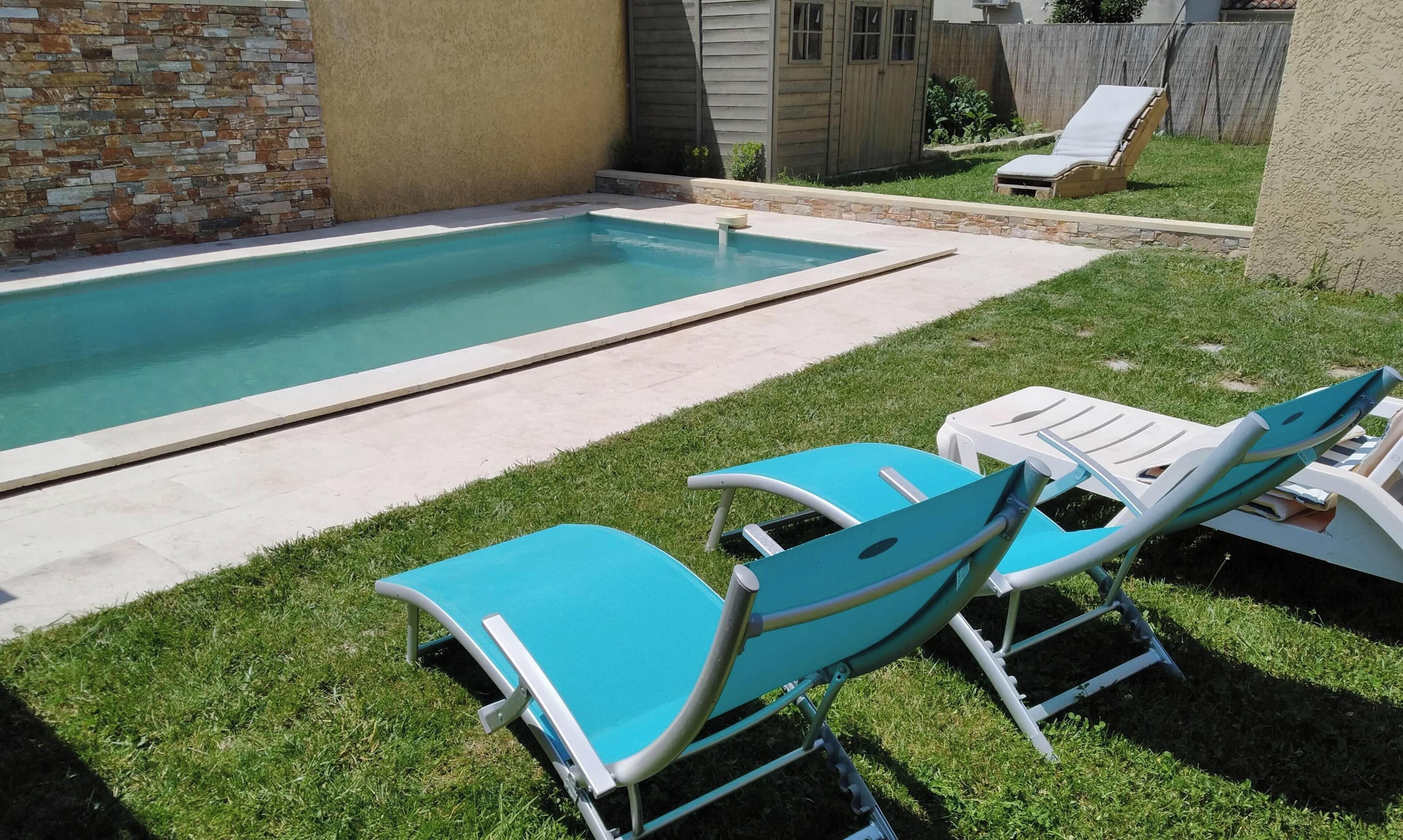 A backyard with a swimming pool, two blue lounge chairs, and a stone wall.