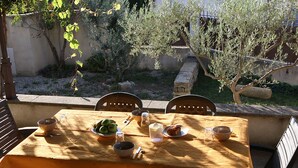 Mesa de comedor al aire libre con mantel, rodeada de sillas, y un jardín y un muro de piedra al fondo.