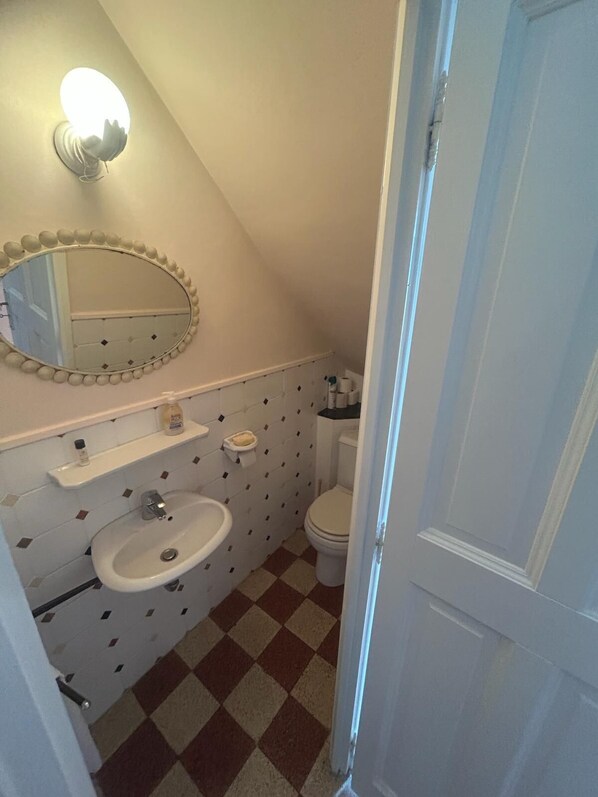 Ein kleines Badezimmer mit einem weißen Waschbecken, Spiegel und Toilette. Der Boden hat ein Schachbrettmuster, und über dem Waschbecken befindet sich ein Handtuchhalter.