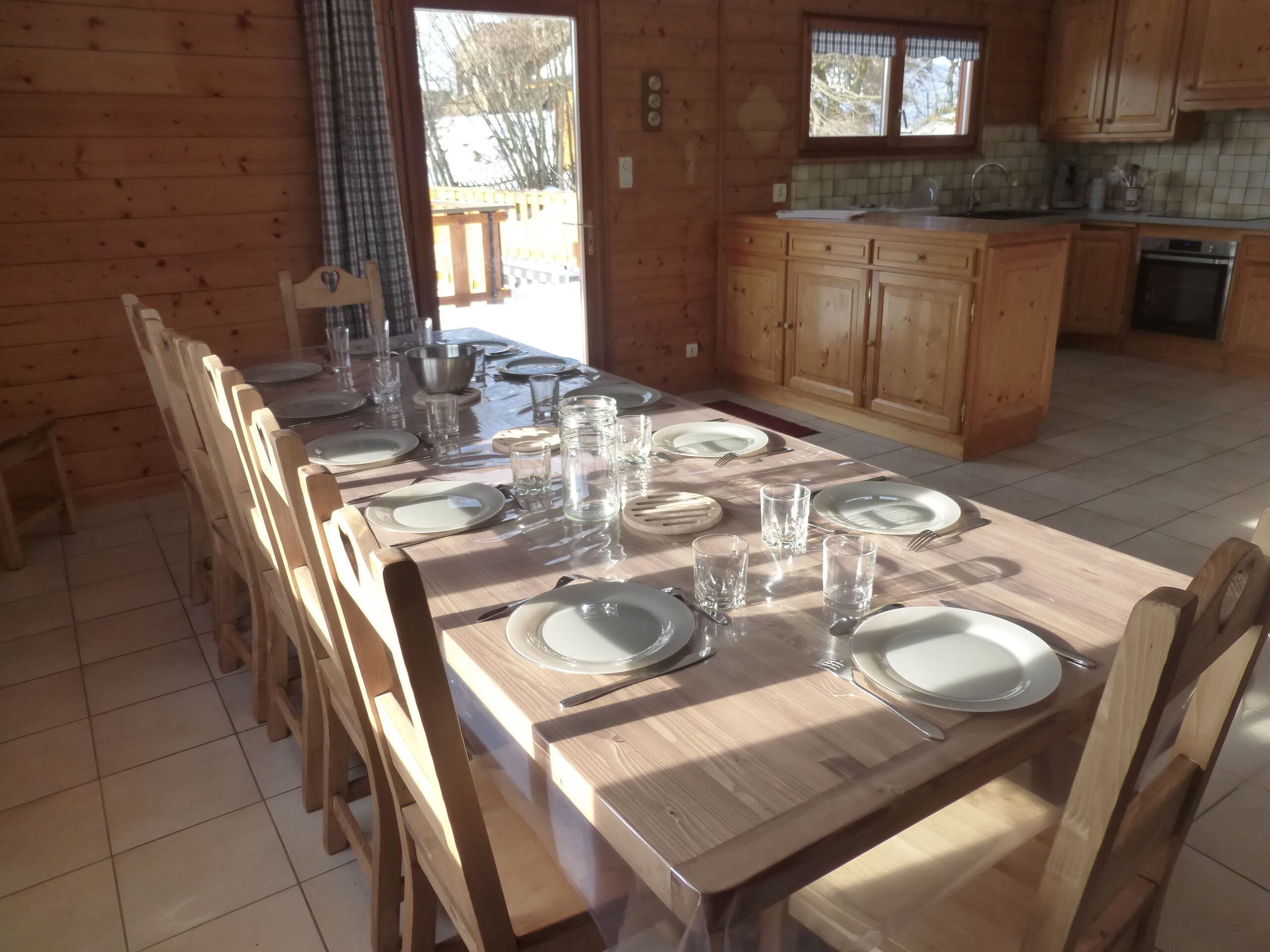Une table à manger en bois, accompagnée de six chaises, un coin cuisine avec des placards en bois et une fenêtre avec des rideaux.