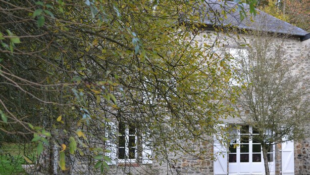 A stone building with white shutters, surrounded by trees and a grassy area. - Cottage with a pond on the border of Brittany and Normandy (La Baconnière)