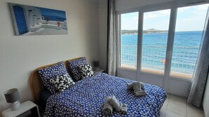 Een slaapkamer met een bed, een plafondventilator, een nachtkastje, een schilderij en een groot raam met uitzicht op de zee.