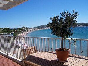 Un balcon avec une table en bois et un olivier en pot, donnant sur une plage animée et un ciel bleu dégagé.