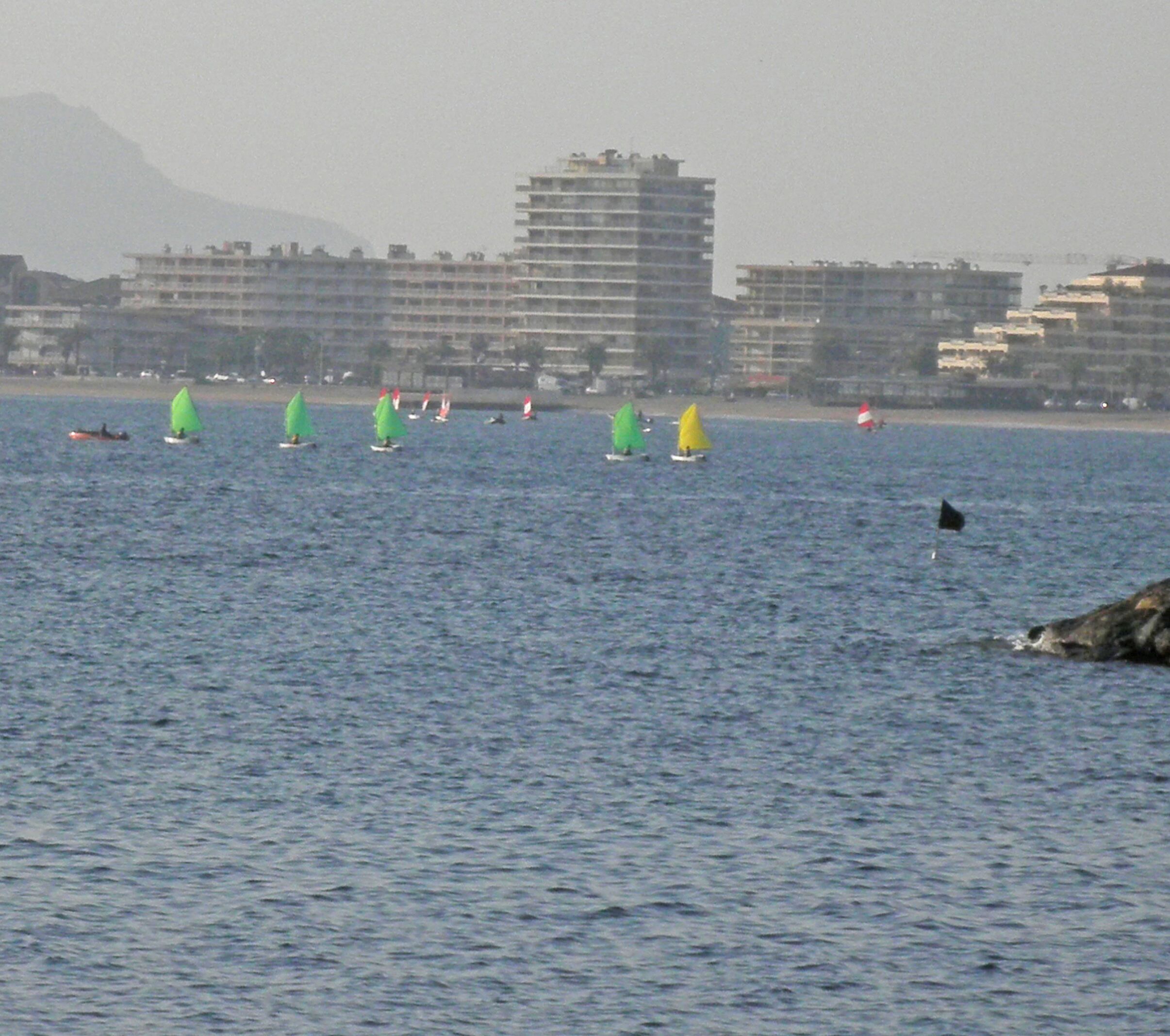 A view of a body of water with several sailboats, a distant mountain, and buildings in the background.