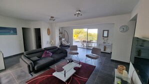 A modern living room with a black sofa, glass coffee table, and two chairs. There is a dining area with a table and chairs visible through a glass door. - Vista Para o Mar, Calmo / sem Vis-à-vis, a 5min da Praia, Piscina com Portões (Calvi)