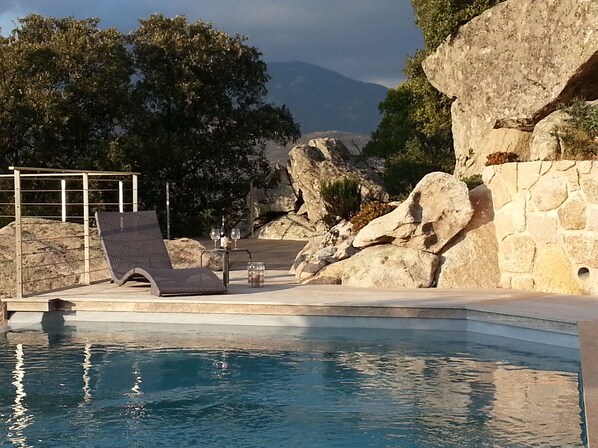 Un área de piscina con una silla de descanso, un muro de piedra y vistas a montañas.