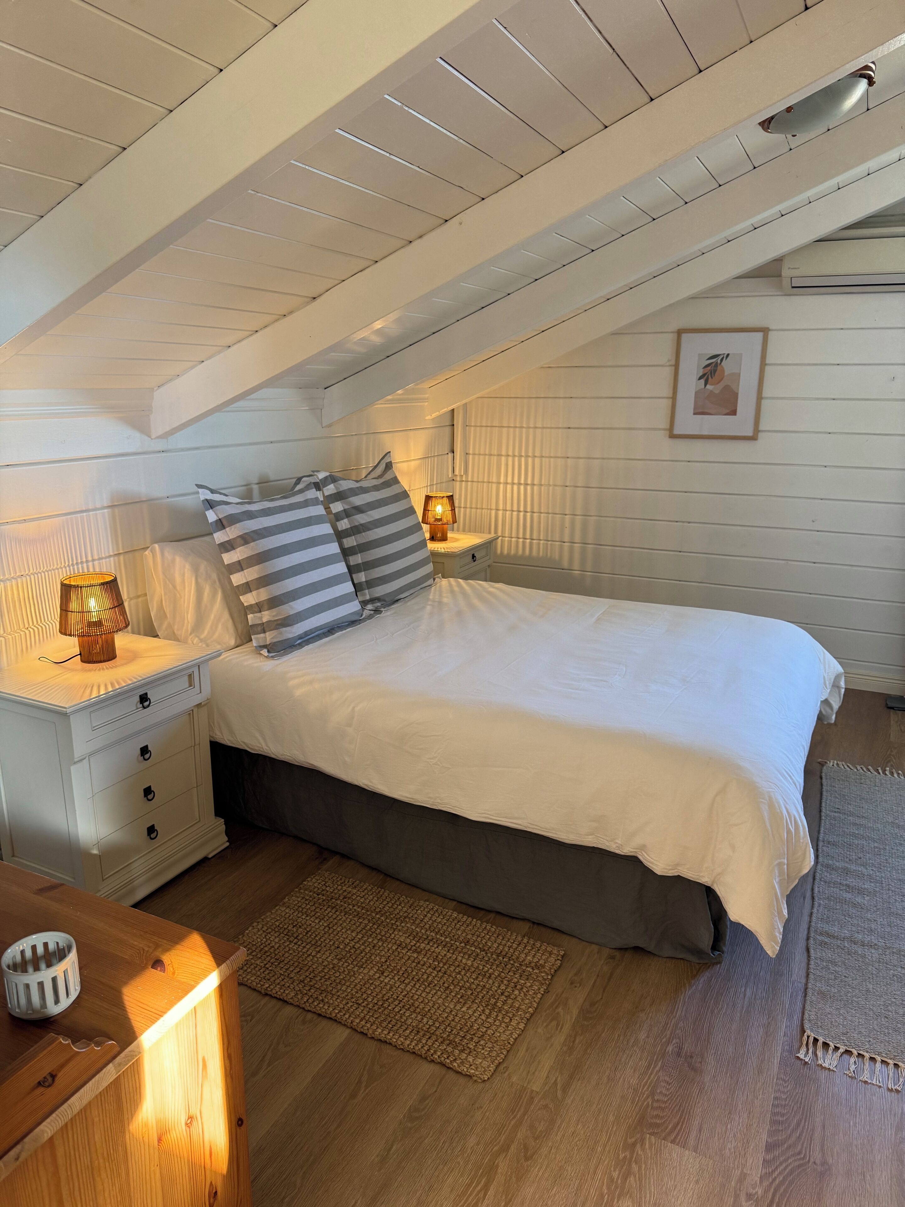 Une chambre avec un plafond en pente, un lit avec une literie blanche, des oreillers à rayures, deux lampes de chevet, une petite table de chevet et un tableau encadré au mur.