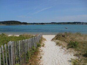 En sandig strandpromenad som leder till ett lugnt hav, med ett trästaket på ena sidan och en liten båt i vattnet.