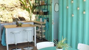 Un espace extérieur abrité, aménagé pour les repas, avec une table turquoise et des chaises blanches, un plafond en bois et un coin cuisine avec un mur bleu et des plantes en pot.