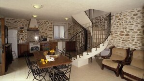 Cocina compacta con pared de piedra, zona de comedor con mesa y sillas de madera, y escalera de caracol que sube a un nivel superior.