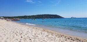 Ein Strand mit klarem, blauem Wasser, Booten und einer hügeligen Landschaft im Hintergrund.