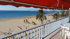 A beach view from a balcony with a red canopy, palm trees, and a playground. - ROSES: Super Apartment on the FIRST LINE OF THE SEA for 6–8 PEOPLE in the CITY CENTER (ROSAS)