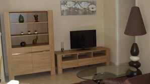 Un salon comprenant un meuble de rangement en bois pour l’équipement audiovisuel, un téléviseur à écran plat, une table basse en verre, un canapé marron et une lampe.