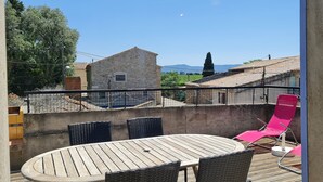 Un balcon doté d’une table ronde en bois et de deux chaises en osier, offrant une vue sur un bâtiment en pierre et un paysage montagneux.