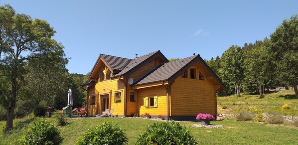 A two-story wooden house with a sloped roof, surrounded by greenery and trees.