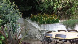 Un patio con una mesa redonda y sillas, rodeado de plantas y un muro de piedra.