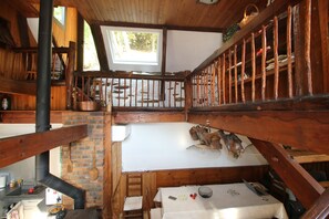 Interior rústico de una cabaña con cama en un altillo, vigas de madera, chimenea de ladrillo y tragaluz.