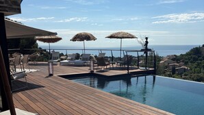 Terraza de madera con sillones de descanso y sombrillas con vistas a una piscina y al mar.