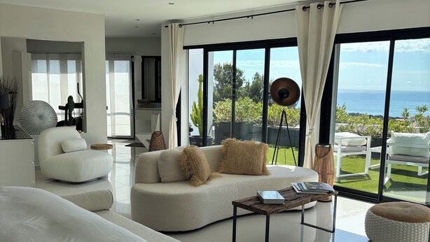Un salón moderno y espacioso con una cama grande, un sofá y una mesa de centro. La habitación tiene ventanas amplias que ofrecen vistas al océano y al follaje.