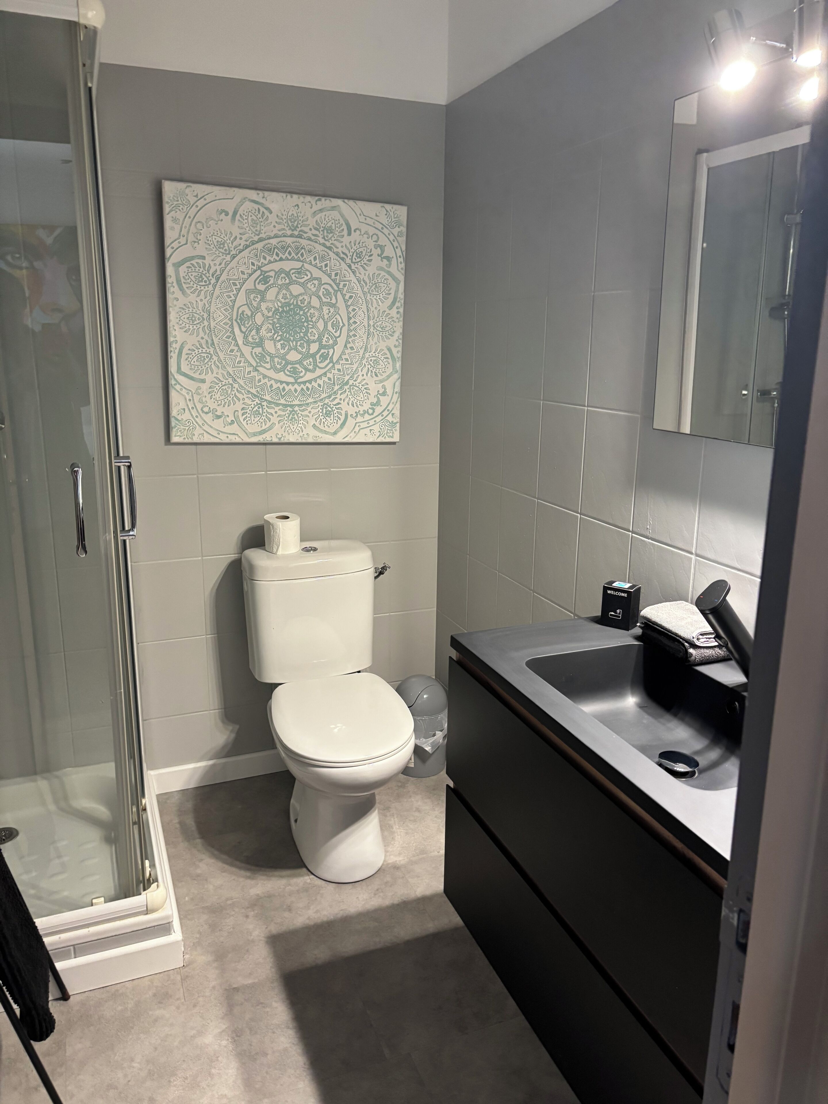 Ein kleines Badezimmer mit Duschkabine aus Glas, Toilette und Waschbecken, versehen mit einem Handtuch und einer mit 'WILLKOMMEN' beschrifteten Schachtel.