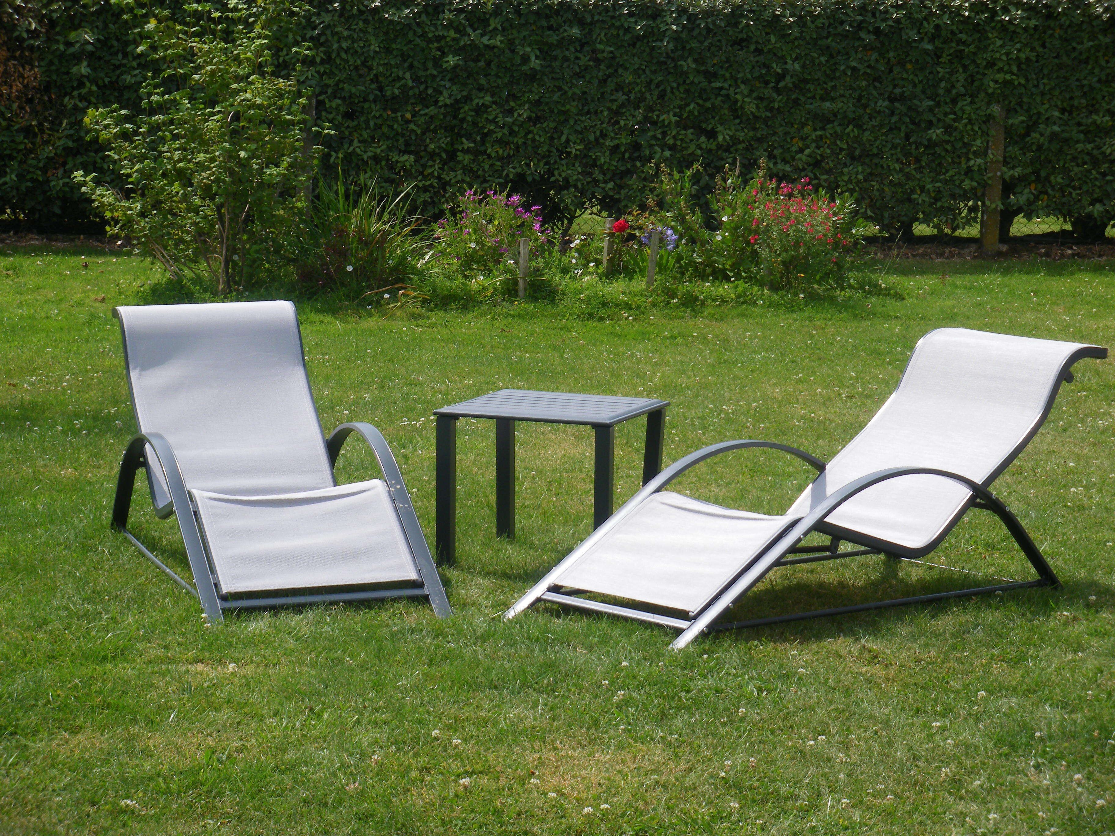 Zwei Lounge-Sessel und ein kleiner Tisch in einer grünen Außenanlage mit Hecke und Blumen im Hintergrund.