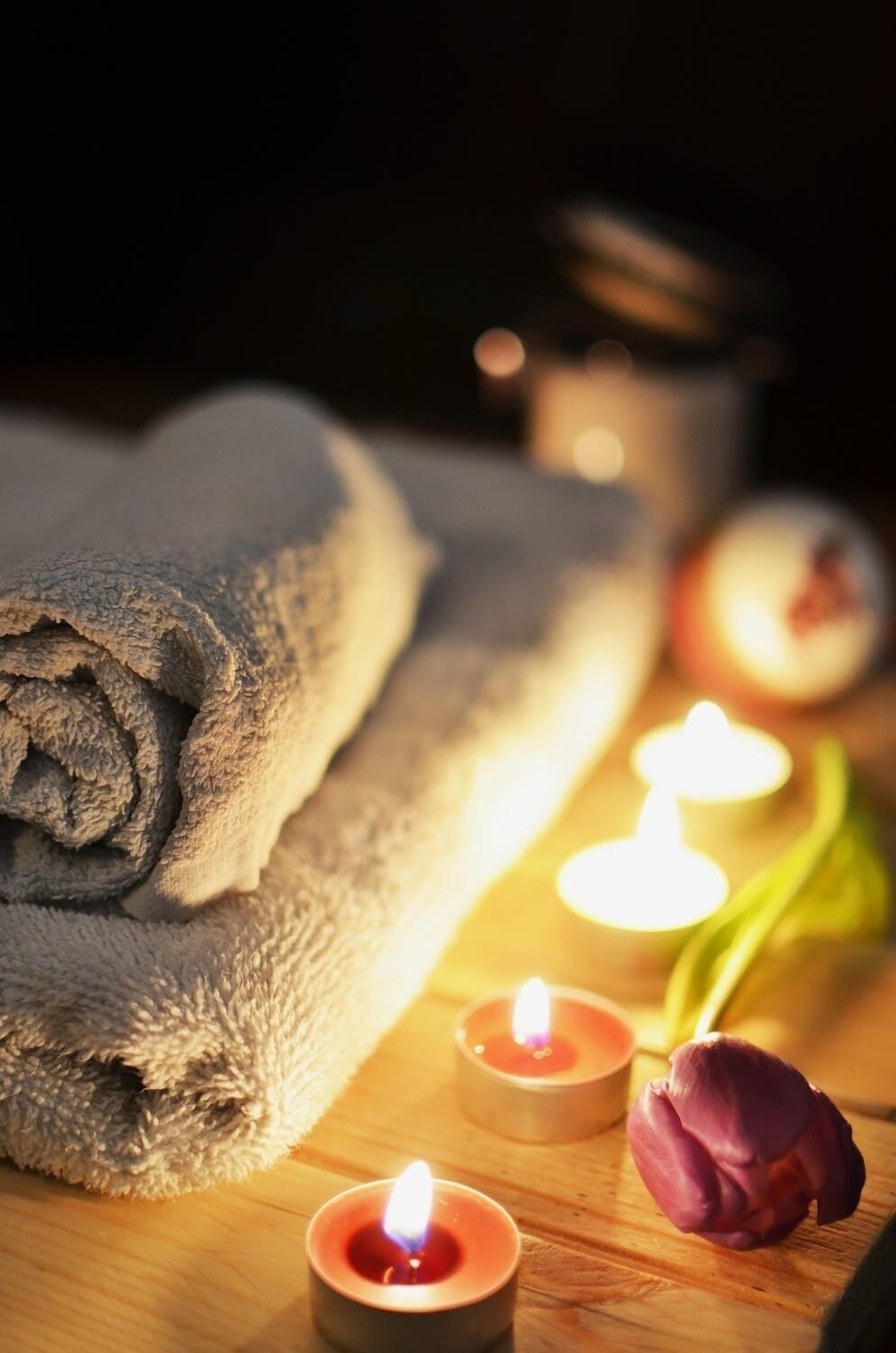 Una superficie de madera con una toalla gris enrollada, dos velas encendidas y una flor morada.