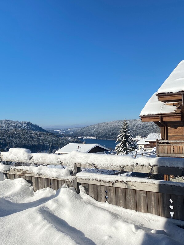 A wooden cabin with a snow-covered roof, surrounded by a wooden fence, overlooking a snowy landscape with distant buildings and a lake.
