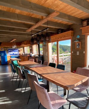 Un intérieur rustique en bois, avec une longue table et des chaises, un poêle et une vue sur l’extérieur.