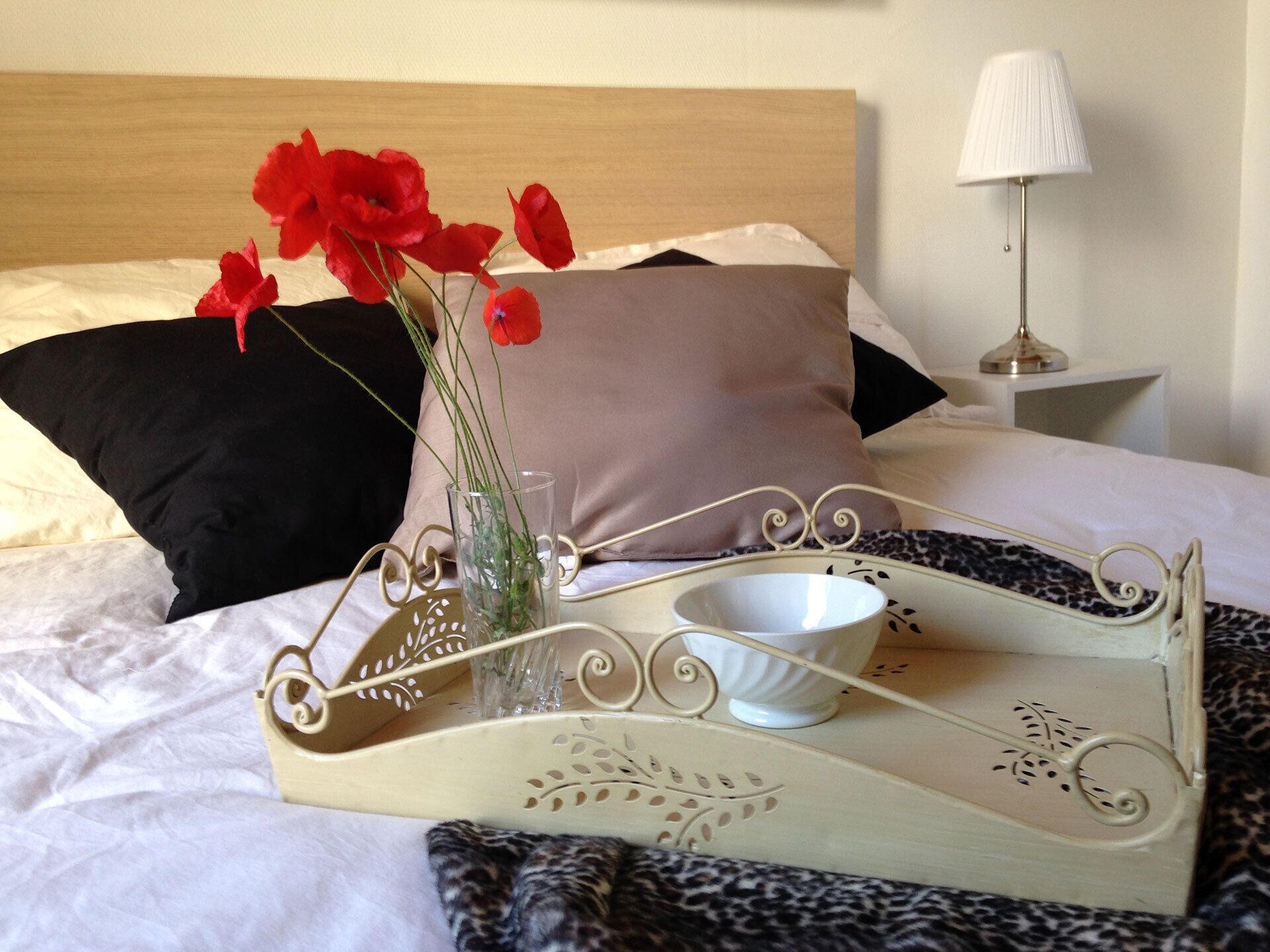Een bed met een dienblad waarop een vaas met rode bloemen, een witte beker en een decoratief dienblad met bloemmotieven staan.