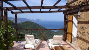 A covered outdoor seating area with two white lounge chairs and a wicker chair with a pink cushion, overlooking a mountainous landscape and the sea. - Traditional house, spectacular view of the Gulf of Porto (Partinello)