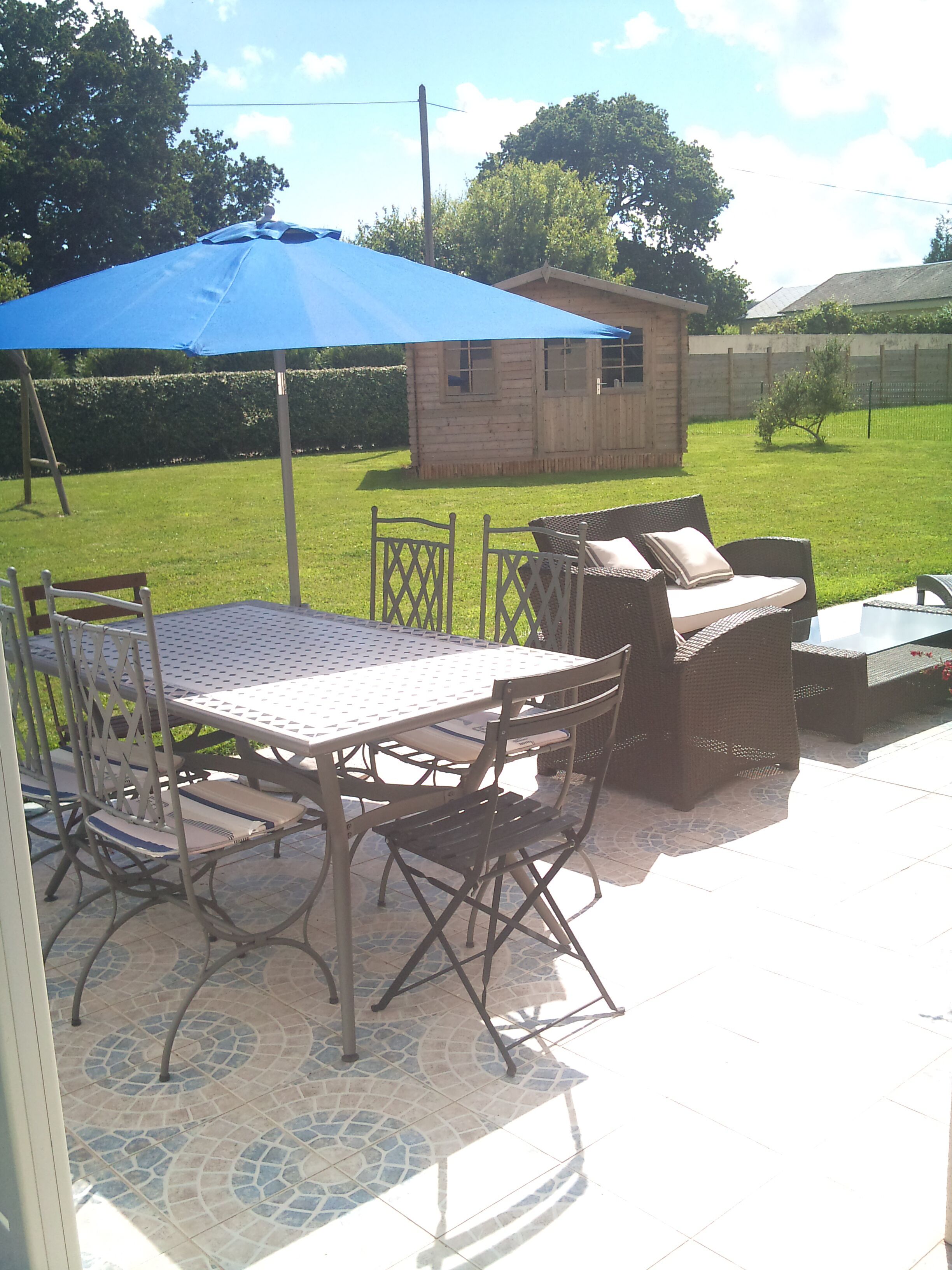 Une terrasse extérieure avec une table et des chaises, un parasol bleu et une petite structure en bois en arrière-plan.