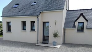 Un bâtiment à deux étages, avec un toit gris, une petite entrée dotée d’une porte et un pot de fleurs placé à l’extérieur.