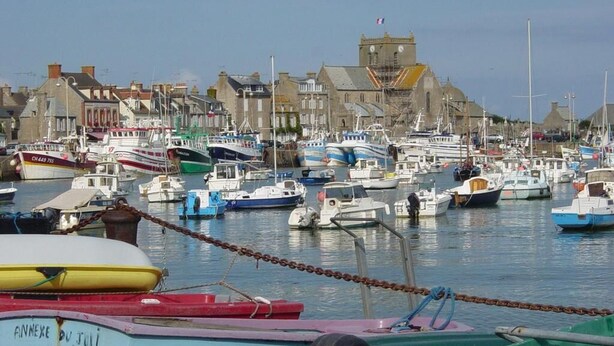 Un port où de nombreux bateaux sont amarrés, un bâtiment imposant doté d’une tour, et un ciel dégagé.