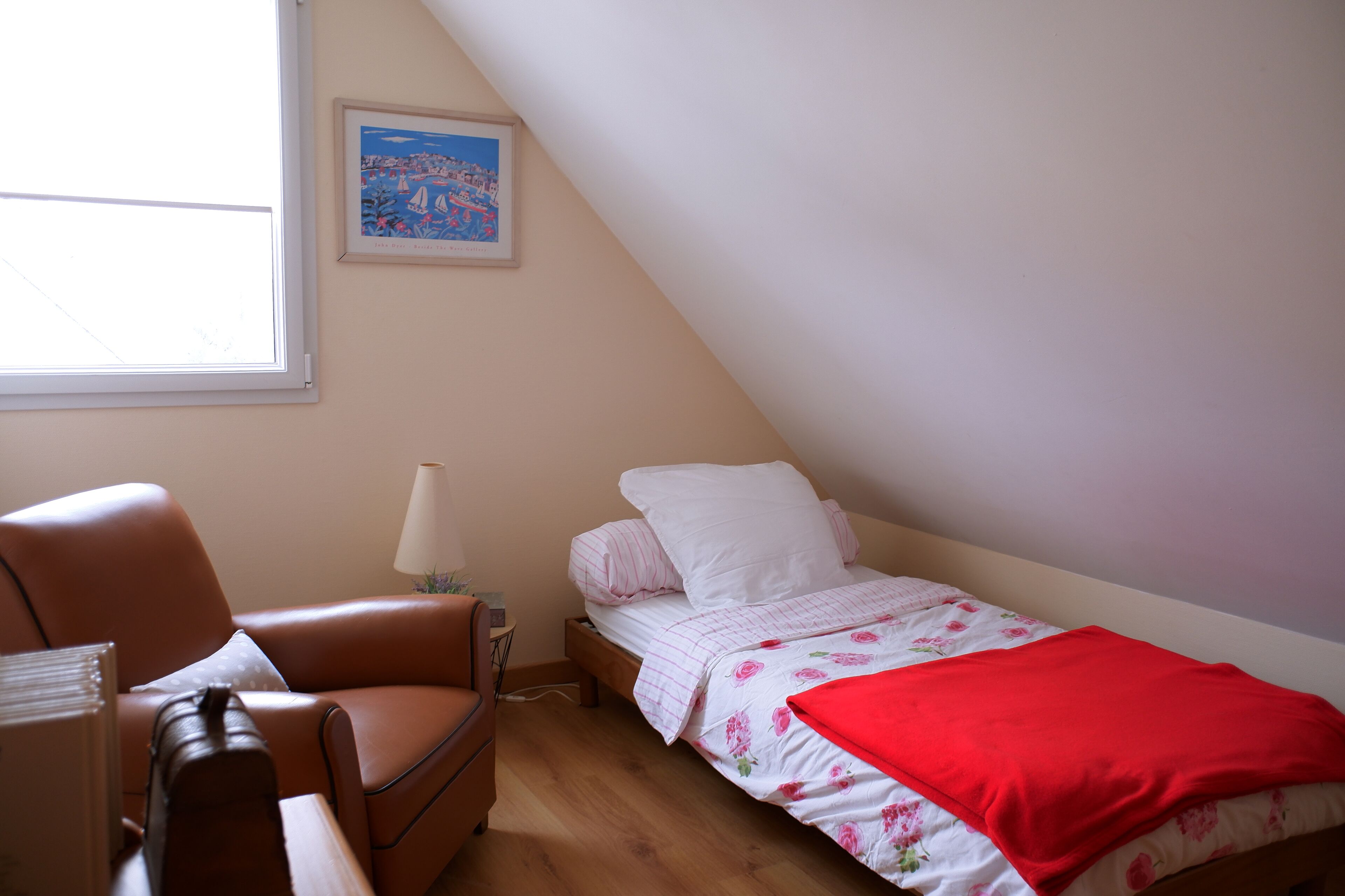 Een kleine, goed verlichte slaapkamer met een enkel bed, een stoel, een lamp en een lijst met een afbeelding aan de muur.