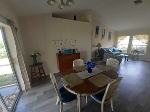 Un comedor con una mesa de madera, sillas azules y un jarrón de flores. Se ve un sofá y una mesita con una lámpara al fondo.