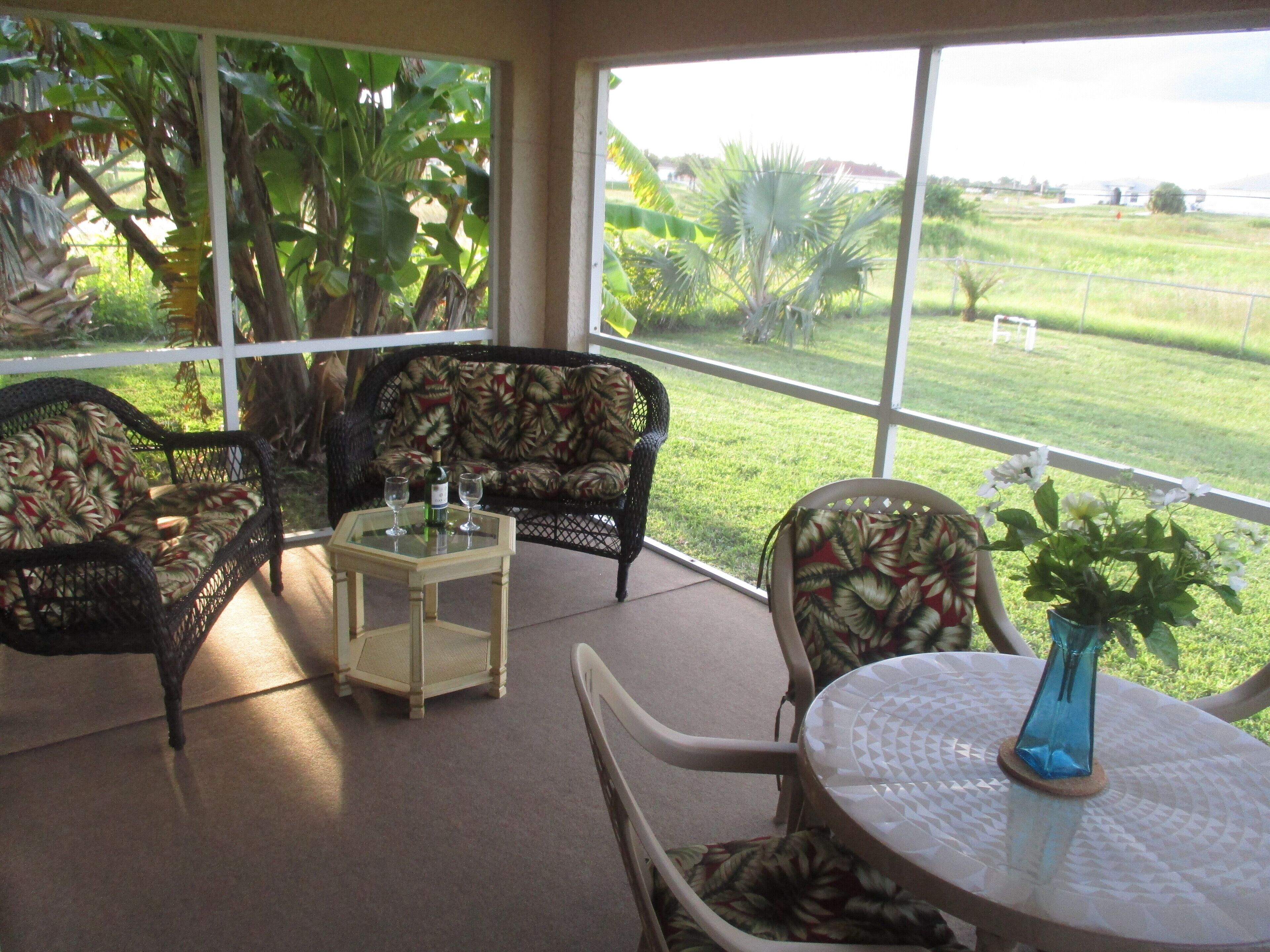 A covered patio with wicker furniture, a glass table, and a view of a grassy field.