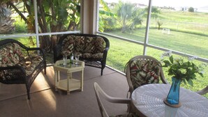 Une terrasse couverte, aménagée avec des meubles en osier, une table en verre et offrant une vue sur un champ verdoyant.