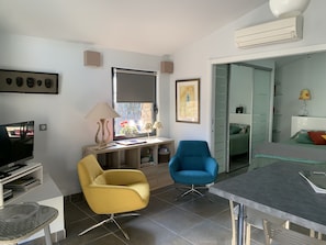 Una sala moderna con comedor, televisor, escritorio con lámpara, una silla azul y una estantería.