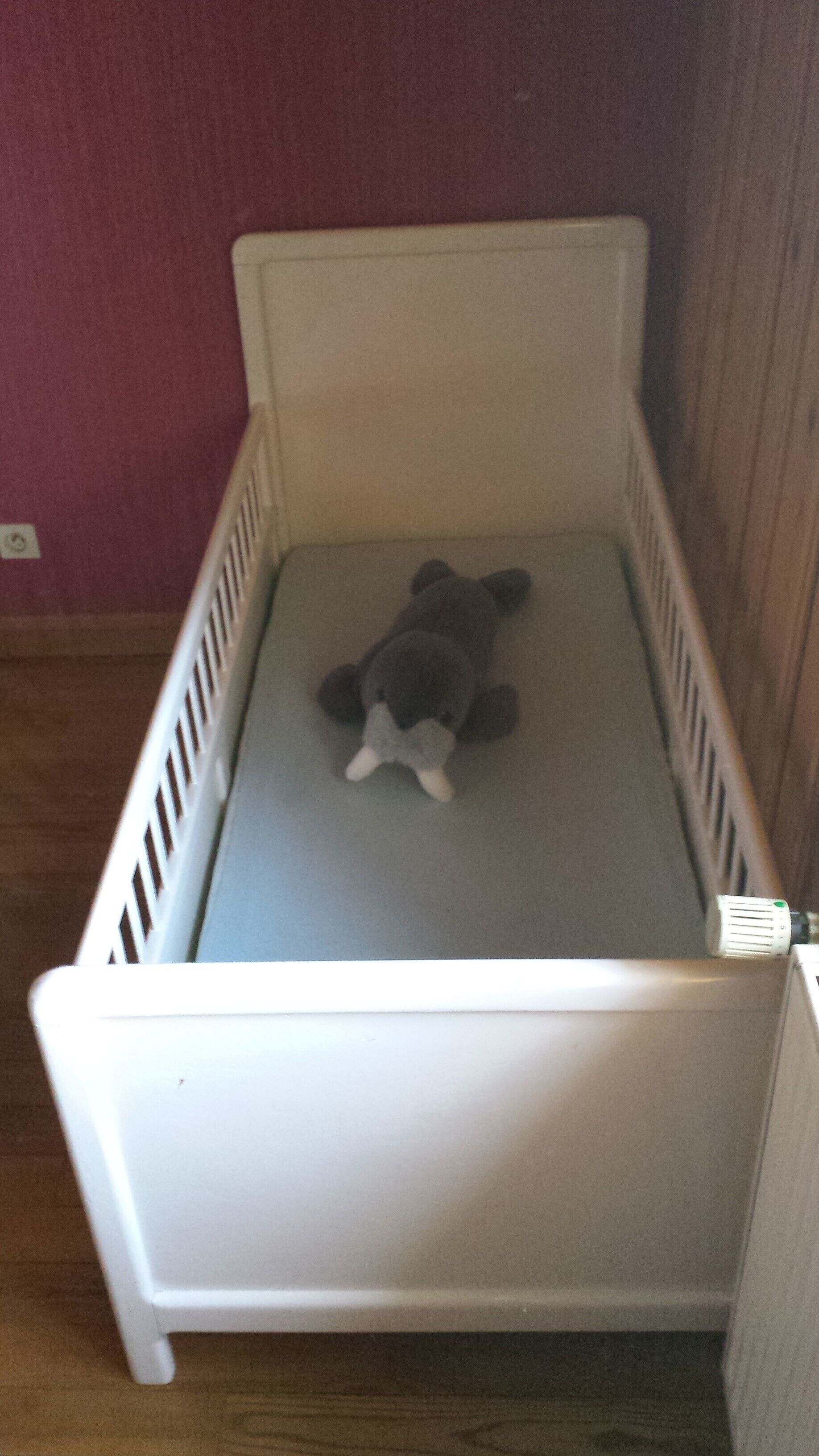 A white crib with a grey mattress, positioned against a wooden wall and a red wall.