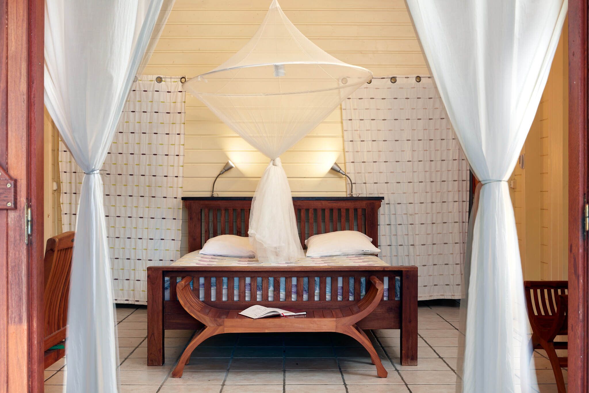 Een houten bed met een hemelbed, witte gordijnen en een betegelde vloer.