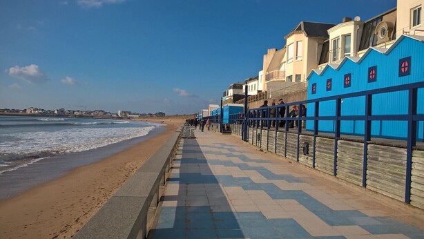 Eine Küstenpromenade mit bepflastertem Gehweg, einem Strand und Gebäuden mit blauen Fassaden.