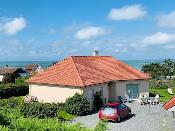 Una casa de un piso con techo de tejas rojas, un porche cubierto con mobiliario exterior y vista al mar.