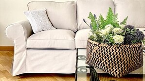 Un sofá color beige con un cojín estampado, una mesa de centro de vidrio con una cesta tejida que contiene plantas, y suelo de madera.