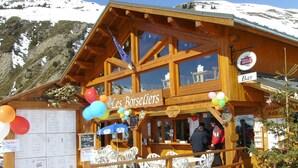 Un chalet en bois avec une enseigne indiquant « Les Borsellers » et une enseigne de bar. On y trouve des chaises et des tables en plastique blanc à l’extérieur.