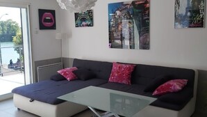 A modern living room with a glass coffee table, a sofa with pink and black cushions, and wall art depicting cityscapes. - Charming cottage by the Seine (Villeneuve-le-Roi)