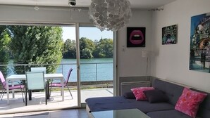 A modern living room with a glass coffee table, a sofa with pink cushions, and a view of a lake through large windows. - Charming cottage by the Seine (Villeneuve-le-Roi)